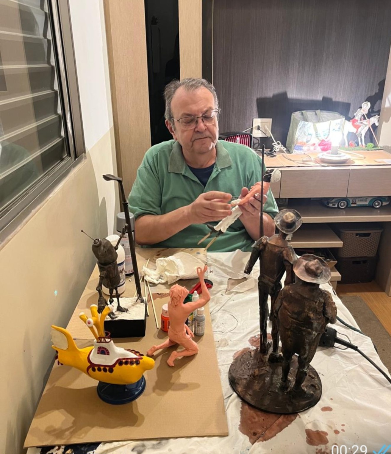 Marcos Prata trabalhando em uma miniatura em sua bancada, cercado por esculturas.